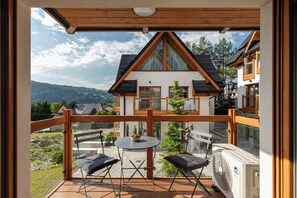 Ein Balkon mit Tisch und Stühlen, einem Ventilator und Blick auf ein Haus mit einem geneigten Dach und Solarmodulen.