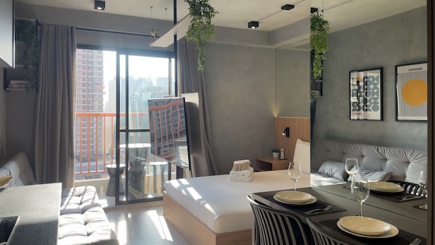 A modern hotel room with a bed, a dining table set for two, a sofa, and a balcony with a view of the city.