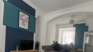 A modern living room with a flat-screen TV on a wooden TV stand, a grey sofa with a blue throw rug, and a large window with white curtains. - Charming Newcastle Home With Garden, Perfect for Families & Contractors (Jarrow)
