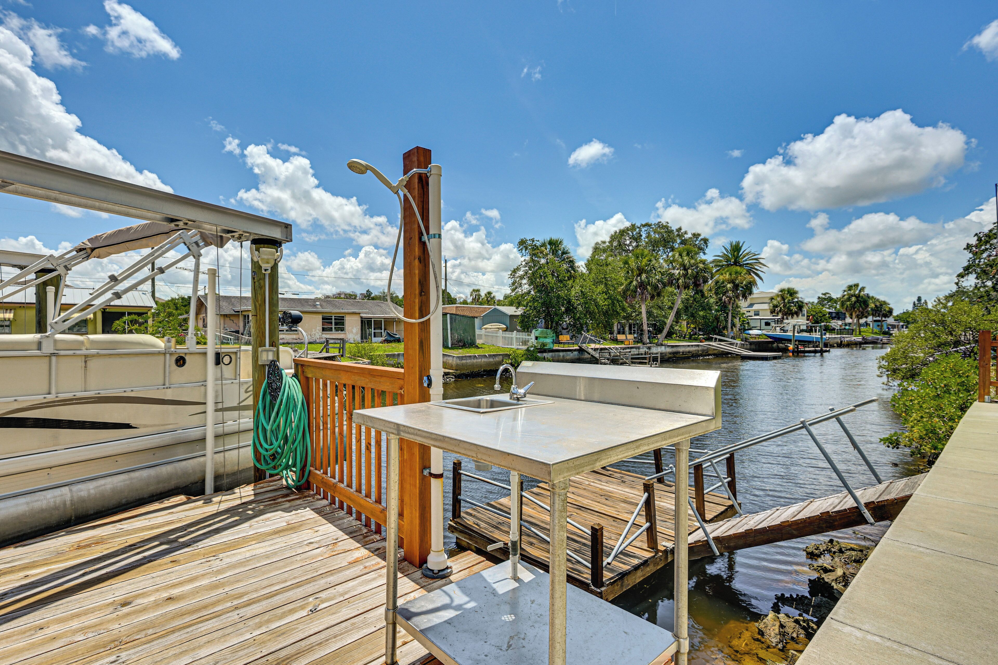 Une terrasse en bois avec une table en métal et un évier, donnant sur une voie navigable paisible, avec des bateaux et des palmiers en arrière-plan.