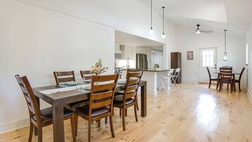 Ampla sala de jantar com mesa e cadeiras de madeira, cozinha ao fundo e teto alto com luminárias pendentes.
