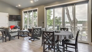 Un amplio comedor con mesa y sillas, ventanas grandes con vistas a árboles y un cuerpo de agua, y un televisor de pantalla plana montado en la pared.