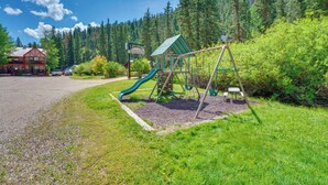 Un terrain de jeux avec un toboggan et des balançoires, entouré d’arbres et avec un bâtiment en arrière-plan.
