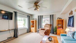 Un salón amplio con un sillón grande, un sofá azul, una mesa de centro de madera y un ventilador de techo. Se observan dos estanterías con diversos objetos y un televisor montado en la pared.