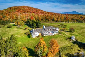 Uma casa grande e com vários andares, com um gramado amplo e montanhas ao fundo.