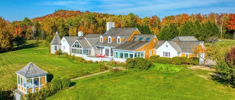 Une grande maison à étages, dotée d’une vaste pelouse devant, d’un belvédère et offrant une vue sur une colline boisée.