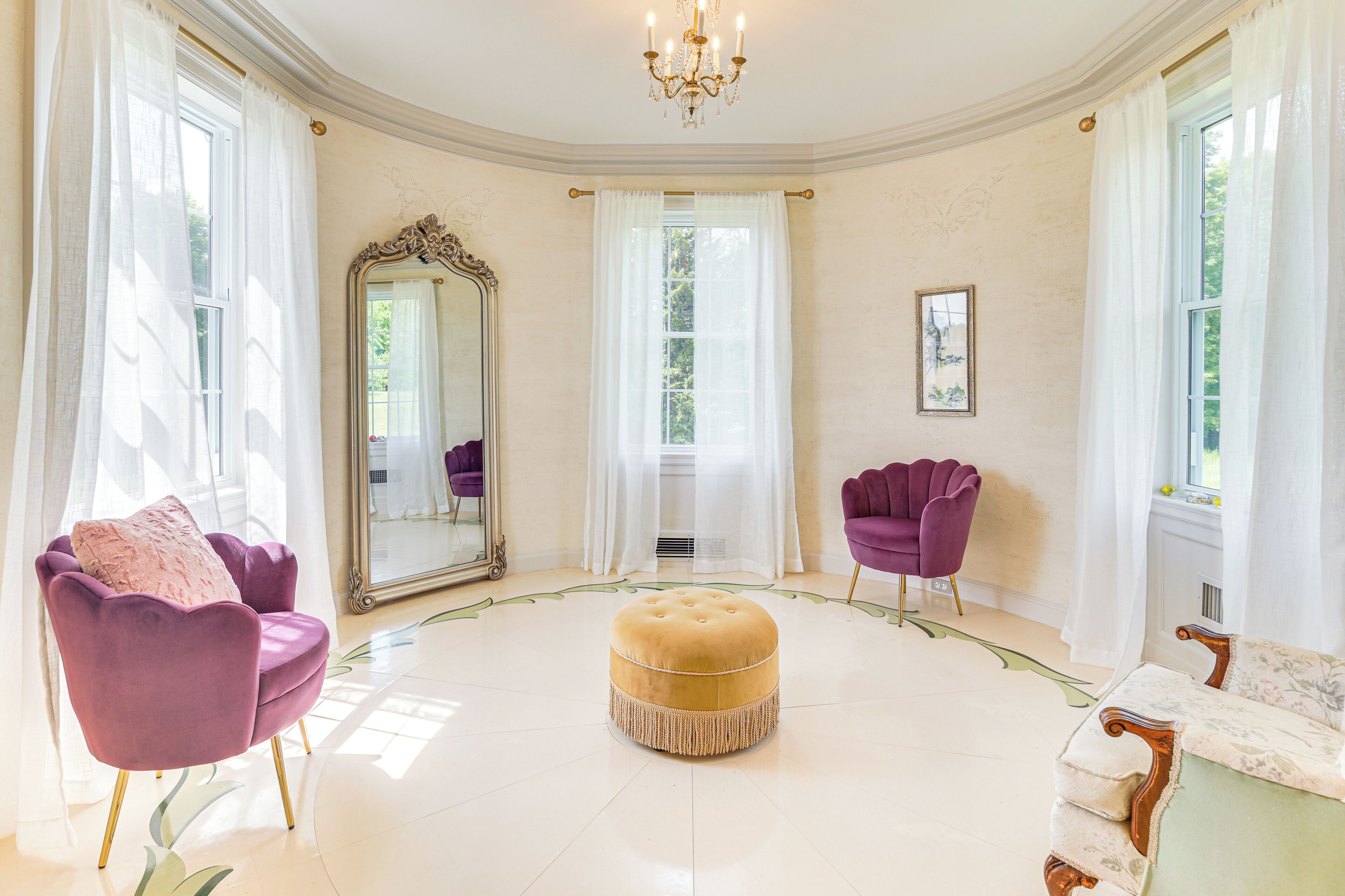 A spacious room with a round ottoman, two purple armchairs, a large mirror, and a chandelier.