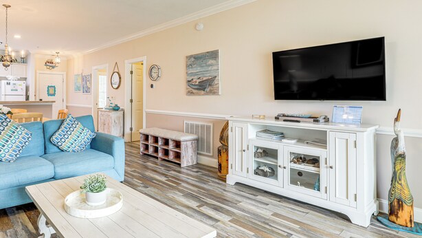 A living room with a blue sofa, a white coffee table with a plant, a white cabinet with glass doors, and a flat-screen TV mounted on the wall. - Golf Course Getaway w/ Pool Access in Myrtle Beach (Myrtle Beach)