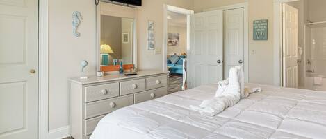 A bedroom with a large bed, a dresser, a TV mounted on the wall, and a bathroom visible through an open door.