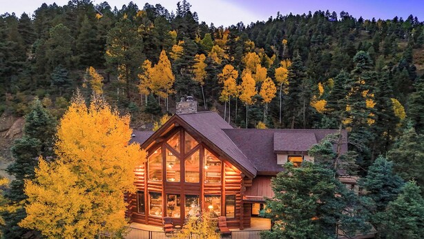 A log cabin with a large glass front, surrounded by autumn foliage and a forested hillside.