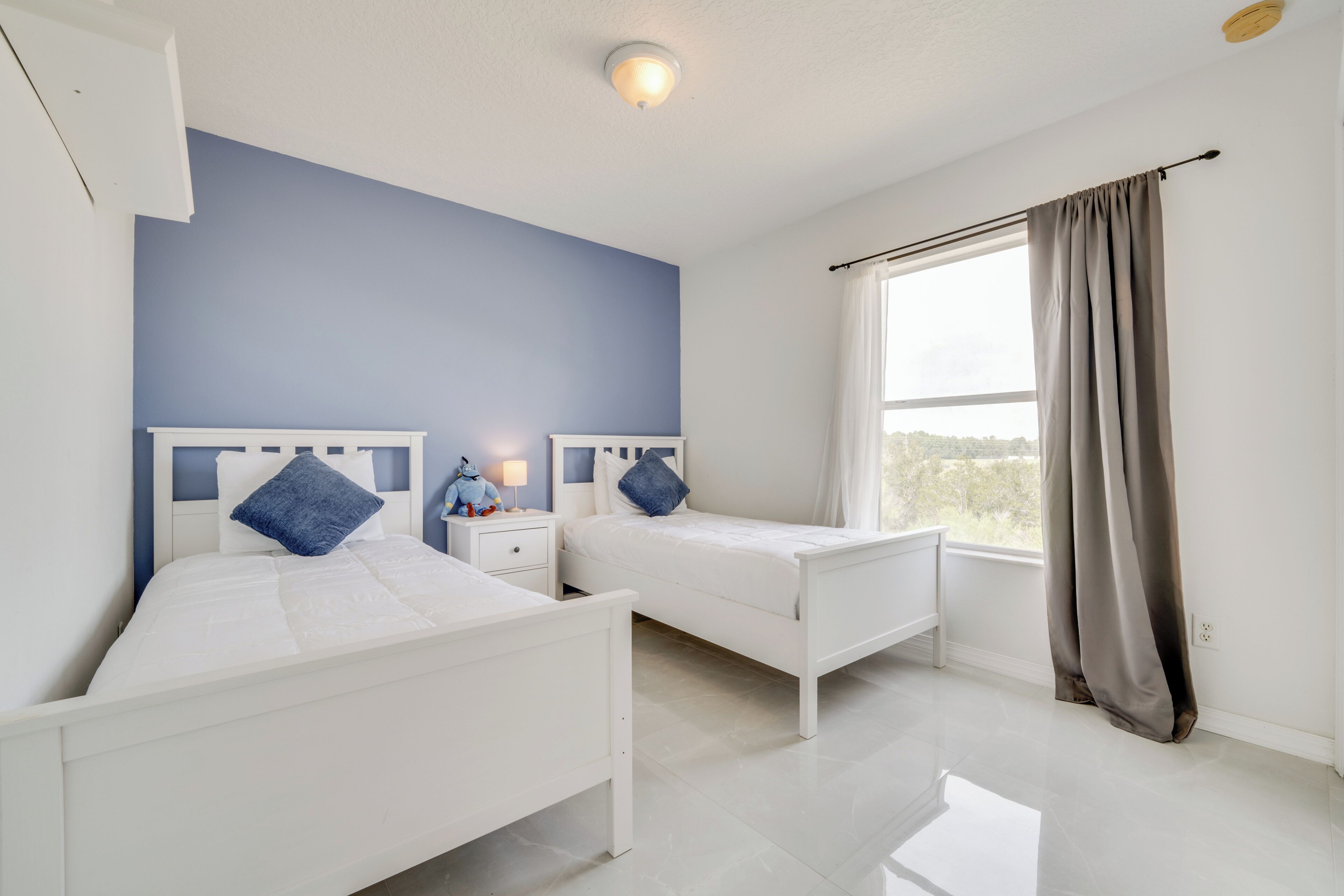 A bedroom with two white beds, a nightstand with a lamp, and a window with curtains.