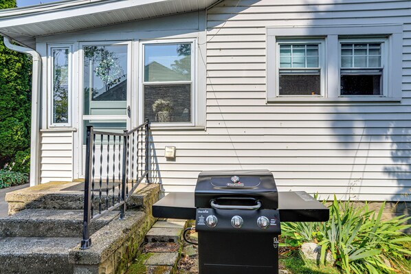 A house with a barbecue grill in front, steps leading to a door, and a window with blinds.