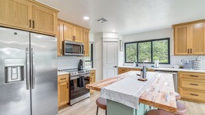 A modern kitchen with wooden cabinets, stainless steel appliances, a large island with stools, and a window providing a view of greenery outside. - Family Fun! Lake Eufaula Home w/ Wraparound Deck (Eufaula)