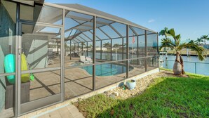 A covered outdoor swimming pool with glass walls, surrounded by a patio area with chairs and a palm tree.