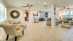 A spacious living room with a beige armchair, a sofa, and a coffee table. There are ceiling fans, recessed lighting, and a large mirror on the wall.