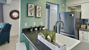 A modern kitchen with teal walls, a stainless steel refrigerator, a sink with a 'Beach House' sign, and a blue chair. - Endless Summer #A30 | Close to Beach + 2 Pools (Panama City Beach)
