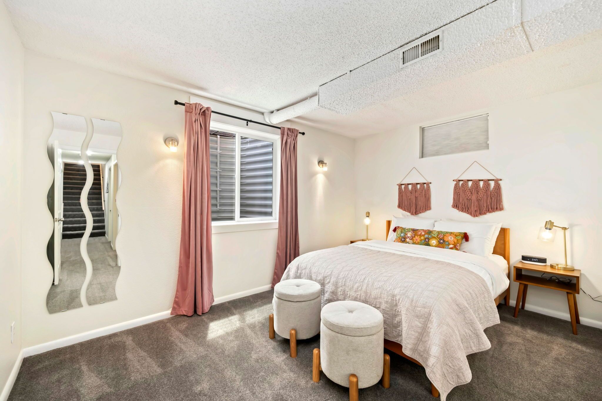 A bedroom with a bed, two ottomans, a nightstand, a mirror, and a window with curtains.