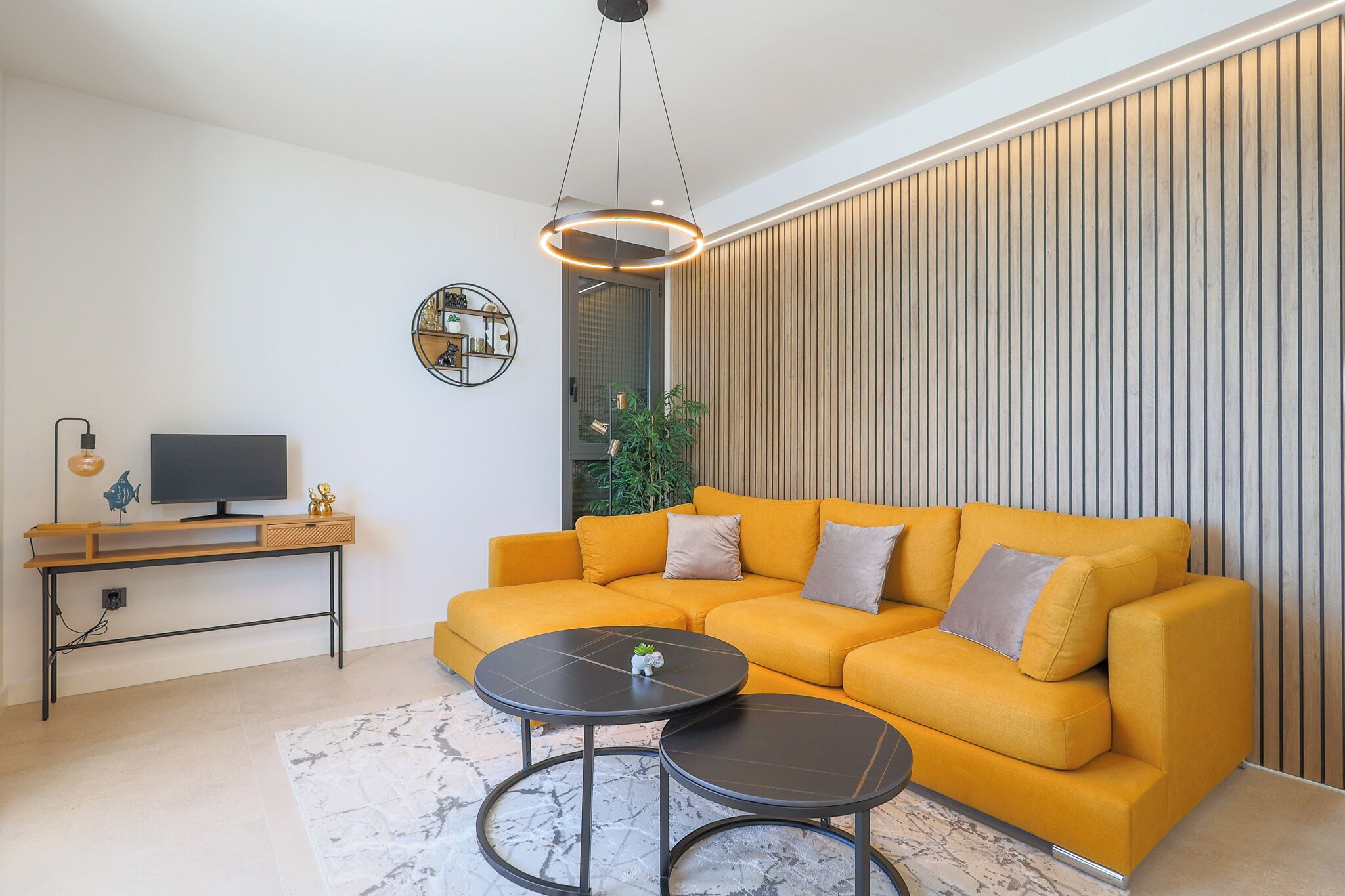 Un salon moderne avec un canapé jaune, deux petites tables rondes noires, un téléviseur posé sur un bureau en bois et un luminaire suspendu.