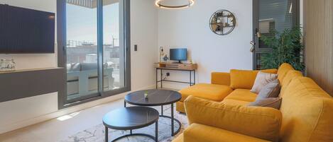 A modern living room with a yellow sofa, two black round tables, a flat-screen TV, and a large sliding glass door leading to an outdoor terrace.
