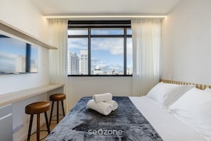 A hotel room with a bed, two bar stools, a window with curtains, and a city view.