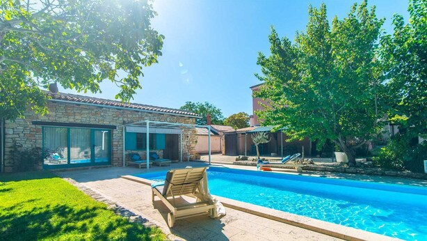 Ein Swimmingpool mit Liegestühlen, ein Steinhaus mit Glasdoor und ein Baum.
