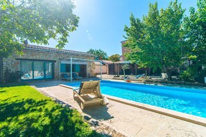 Una piscina con sillas de descanso, un edificio de piedra con puerta de vidrio y un árbol.