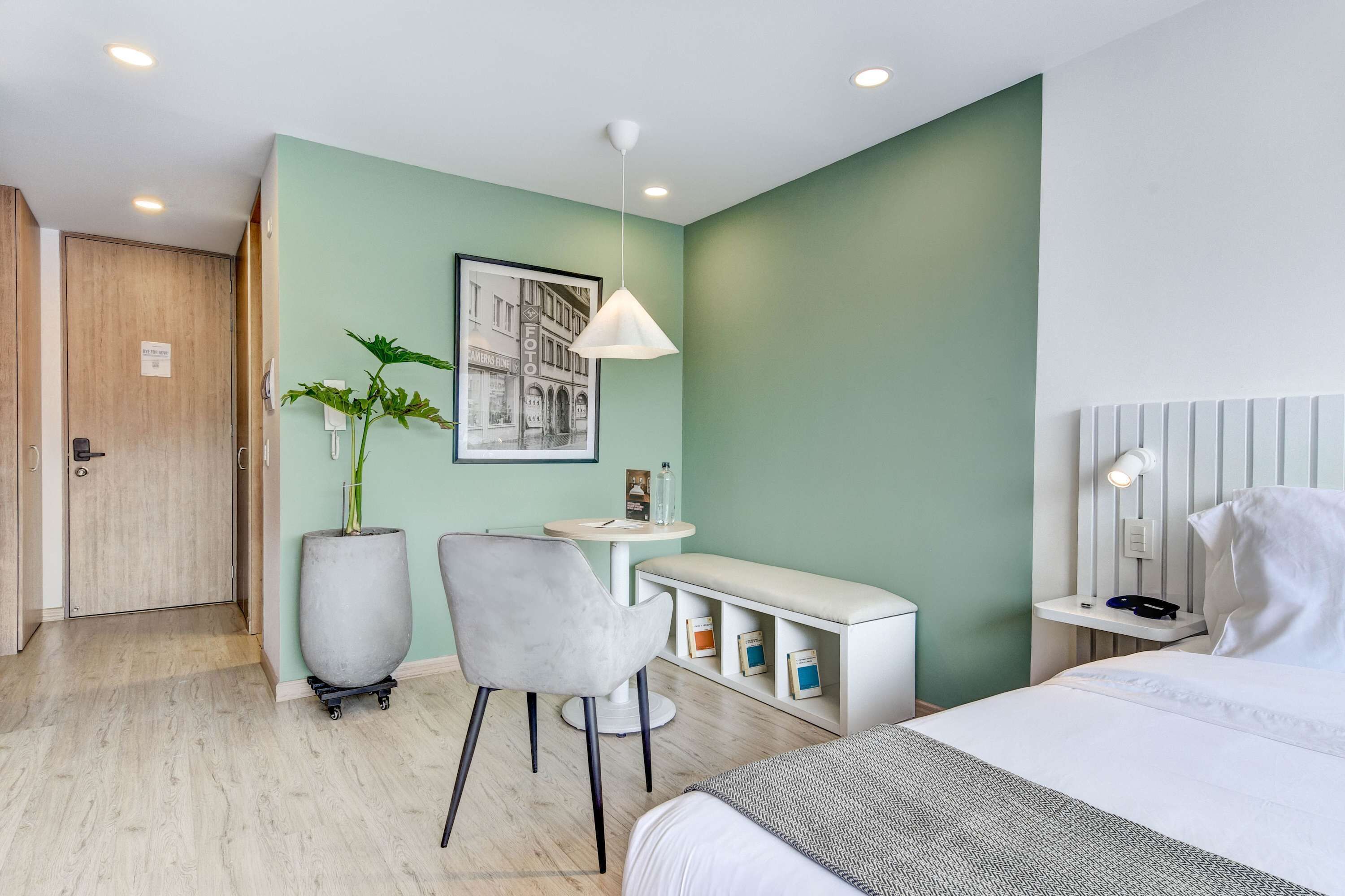 Une chambre d’hôtel comprenant un lit, un bureau, une chaise, une plante en pot, une étagère fixée au mur et une porte.