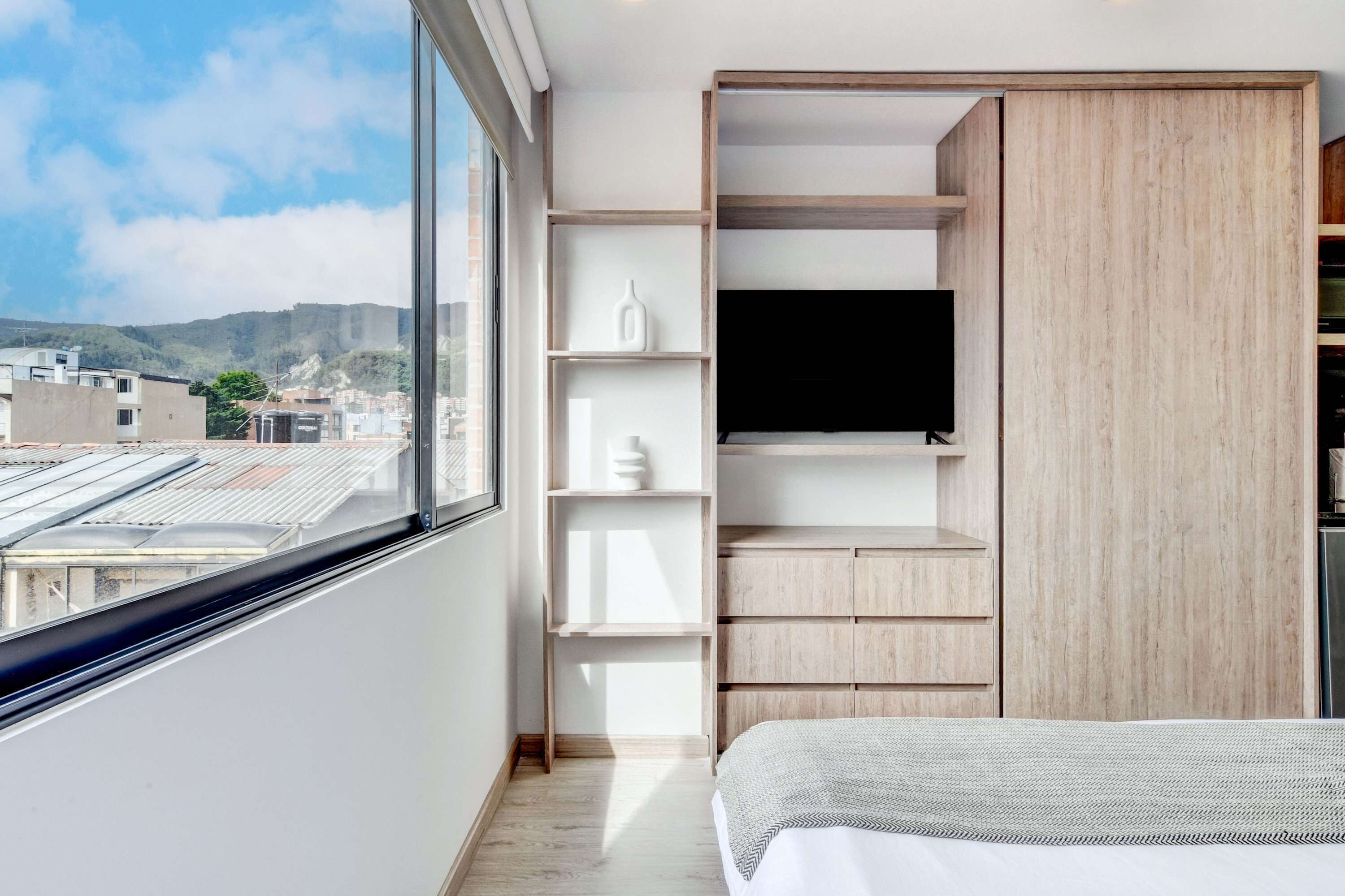 A modern bedroom with a large window, built-in shelving, and a flat-screen TV.