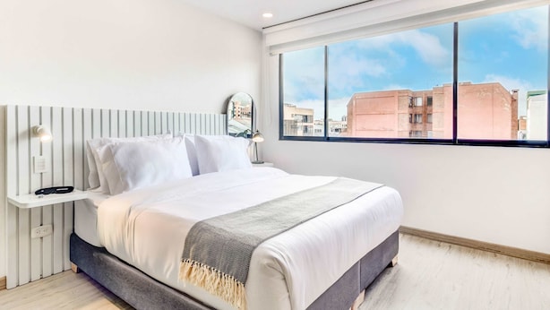 A modern bedroom with a large bed, a bedside table with a lamp, and a view of the city through a large window.