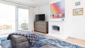 A modern bedroom with a large window, a flat-screen TV, a fireplace, and a painting on the wall. - 3-bed Beach Bungalow Garden 150m to Sea (Chichester)