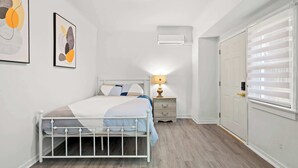 A bedroom with a white metal bed, a bedside table with a lamp, a wall-mounted air conditioner, and a door with blinds.