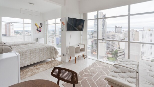 A modern hotel room with a large bed, a desk, and a TV. The room has large windows with a city view. - Studio cond c/ piscina 180m Hard Rock café ESR1912 (Curitiba)