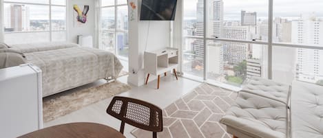 A modern hotel room with a large bed, a desk, and a TV. The room has large windows with a city view.