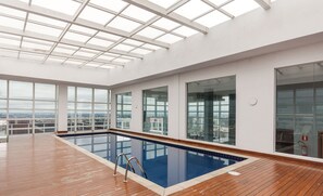 Une piscine intérieure spacieuse avec une terrasse en bois, de grandes fenêtres offrant une vue sur la ville et une verrière.