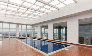 Una piscina interior amplia con cubierta de madera, ventanas grandes con vistas a la ciudad y un tragaluz.