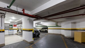Eine Parkgarage mit weißen Wänden, gelb-schwarzen Markierungen und einem grauen Boden. Autos stehen geparkt, ein Einkaufswagen und ein Schild, das einen Ausgang kennzeichnet.