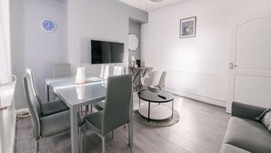 Een moderne woonkamer met een glazen eettafel, grijze stoelen, een ronde salontafel, een flatscreen televisie, een lamp en een bank.