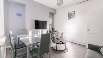 A modern living room with a glass dining table, grey chairs, a round coffee table, a flat-screen TV, a lamp, and a sofa.