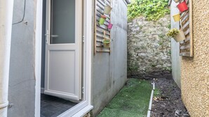 Een smalle buitenruimte met een deur die naar een kamer leidt, een stenen muur en een klein stukje kunstgras.