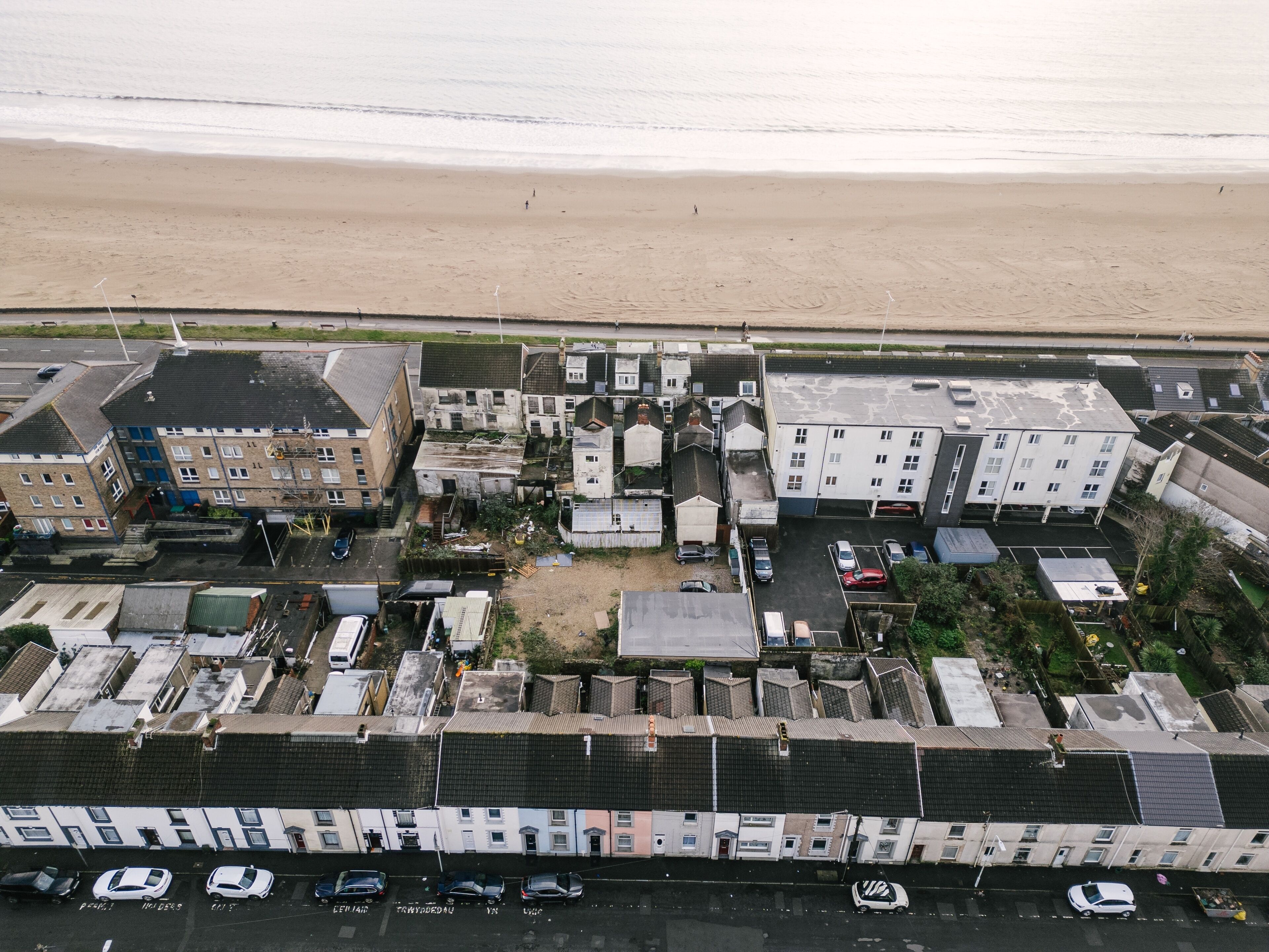 An aerial view of a coastal residential area with buildings, a parking lot, and a beach in the background.