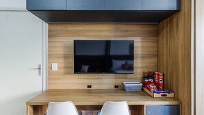 Ein modernes Zimmer mit Schreibtisch aus Holz, zwei weißen Stühlen, einem flachbildfernseher an der Wand und einer kleinen Kommode darunter.