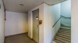 Ein schmaler Flur mit einer Tür, einem an der Wand montierten Gegensprechanlage und einer Treppe mit Metallgeländern.