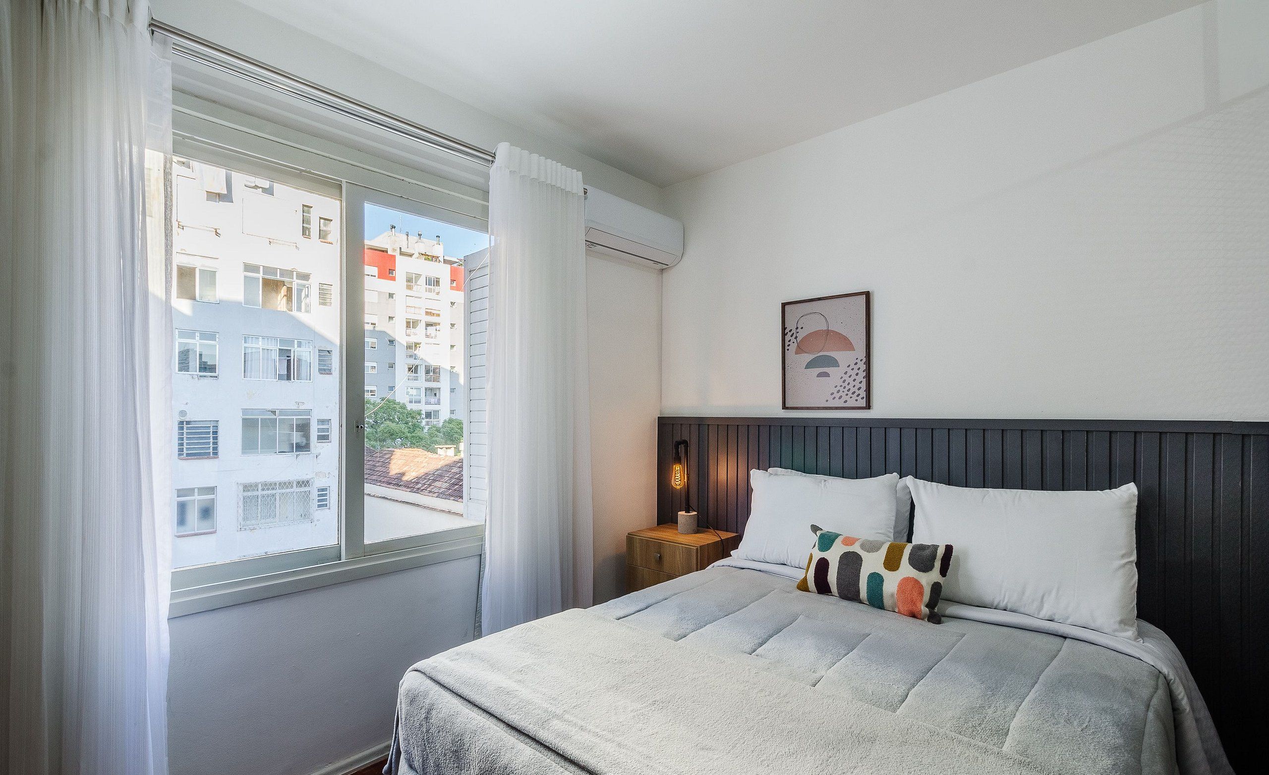 A neatly arranged bedroom with a bed, a bedside table, a wall-mounted lamp, and a window with curtains.