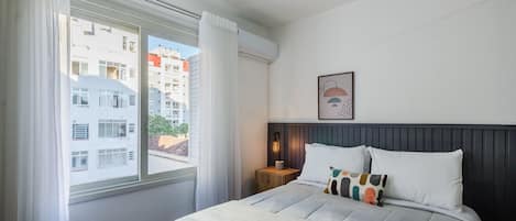 Un dormitorio ordenado con cama, mesita de noche, lámpara de pared y ventana con cortinas.