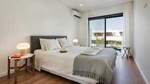 A modern bedroom with a large bed, bedside tables with lamps, a view of buildings through a large window, and an air conditioning unit on the wall.