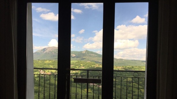 Een balkon met metalen hekken en twee kleine krukjes, met uitzicht op een bergachtig landschap onder een heldere lucht.