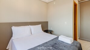A neatly made bed with white linens and a grey blanket, a small round table beside it, and a wooden door leading to another room. - Loft de luxo com Garagem no Petrópolis AHO1402 (Porto Alegre)