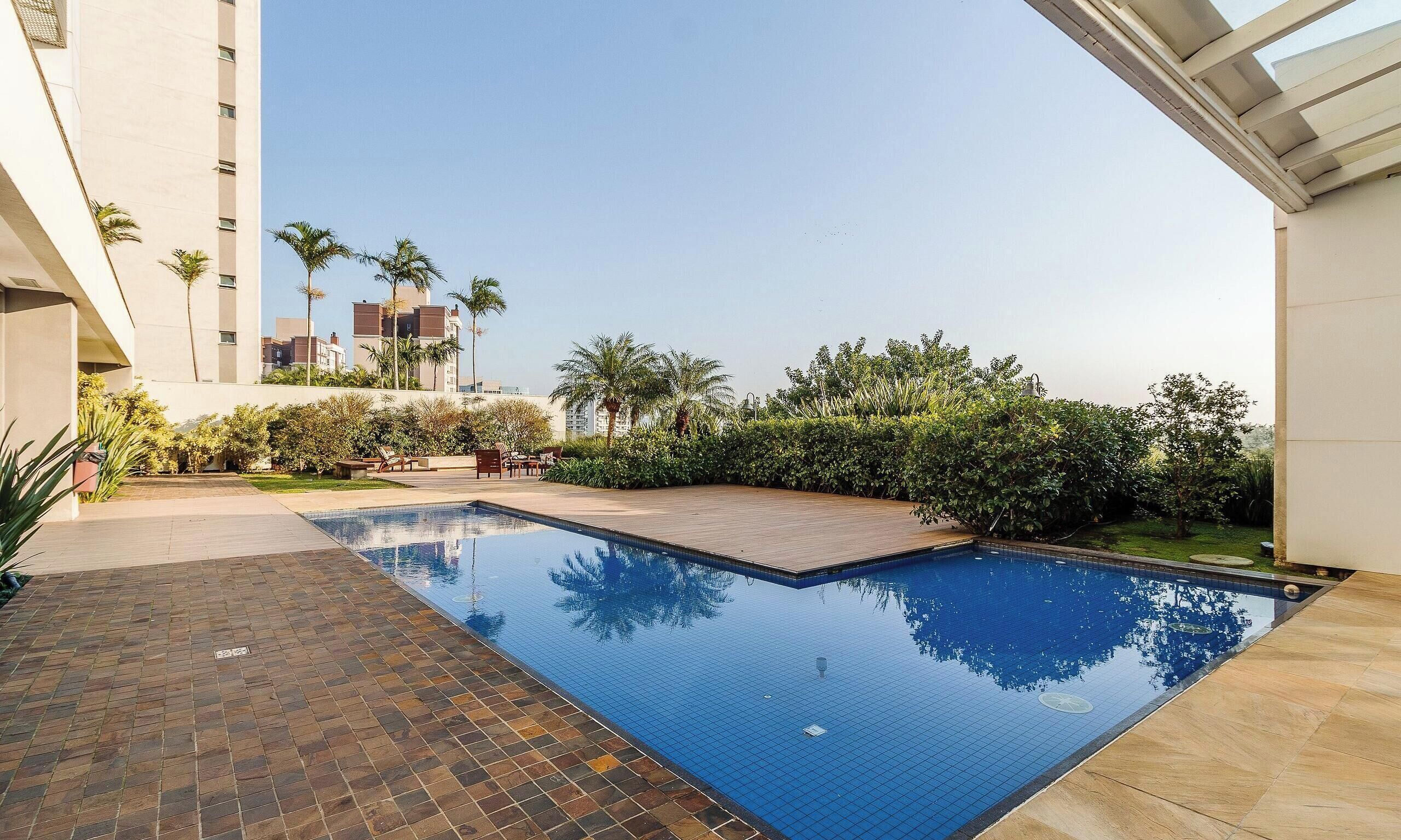 A swimming pool with a tiled deck area, surrounded by greenery and modern buildings in the background.