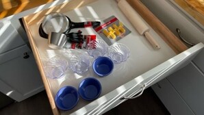 Eine Schublade mit verschiedenen Gegenständen, darunter ein Nudelholz, Scheren, eine Rolle gelbes Klebeband und mehrere durchsichtige Plastikbehälter.
