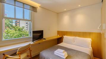 A hotel room with a bed, a desk, a chair, a TV, and a window with a view of greenery.